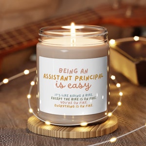 Puede incluir: Una vela encendida en un tarro de vidrio transparente con una etiqueta blanca. La etiqueta dice "BEING AN ASSISTANT PRINCIPAL is easy" con texto adicional debajo. La vela está sobre un pequeño posavasos de madera, rodeada de luces de cadena.