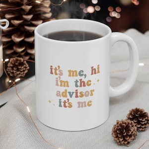 May include: White ceramic mug with a colorful graphic that reads "it's me, hi i'm the advisor it's me". The mug is filled with coffee and has a white handle.