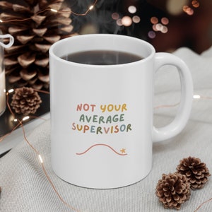 Puede incluir: Taza de café de cerámica blanca con asa negra. La taza tiene el texto "NOT YOUR AVERAGE SUPERVISOR" en letras de colores. El texto está en una fuente caprichosa.
