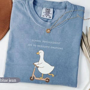 May include: Blue jean colored t-shirt with the text "SCHOOL PSYCHOLOGIST OFF TO REGULATE EMOTIONS" and a graphic of a white duck riding a scooter. A gold necklace with a pendant is draped across the shirt.