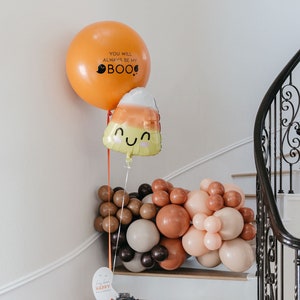 May include: A Halloween-themed balloon arrangement with an orange balloon that says "You will always be my BOO!" and a candy corn shaped balloon. A garland of brown, tan, and peach balloons is on the stairs, with a bucket of treats.