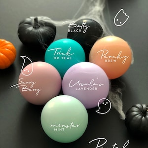 May include: A collection of pastel-colored ghost-shaped ornaments with names like "Trick or Teal", "Peachy Brew", "Scary Berry", "Ursula's Lavender", and "Monster Mint". The ornaments are arranged on a black surface with black pumpkins and spiderwebs.