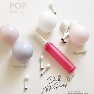 May include: A pink, red, and white double action balloon pump with a white nozzle and a white handle. The pump is surrounded by white, pink, and purple balloons. The text "Double Action Pump" is written in black cursive on a white background. The text "OUR KITS INCLUDE *SINGLE ACTION PUMPS* ONLY" is written in black on a white background.