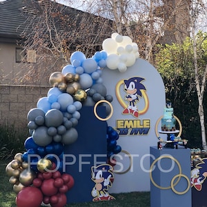 May include: A Sonic the Hedgehog themed birthday party backdrop with blue, gold, and red balloons, a blue and white backdrop with the text "Emile is Five", a blue cake stand with a Sonic the Hedgehog cake, and two blue pedestals with gold rings.