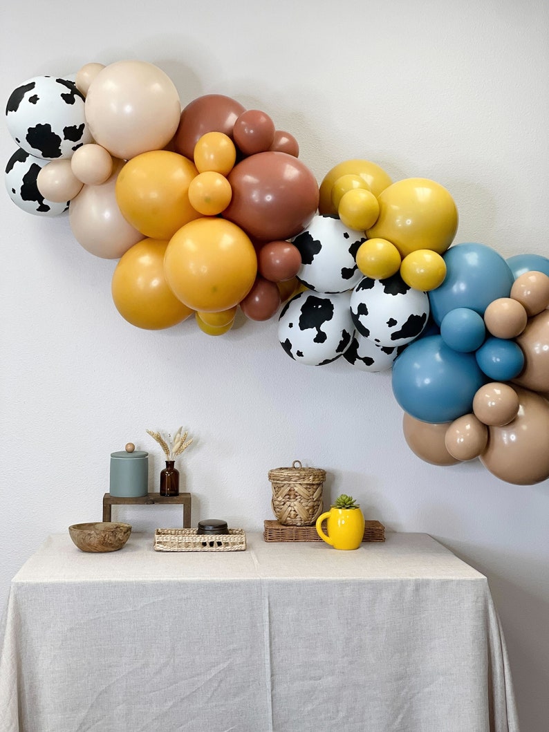 May include: A balloon arch featuring brown, yellow, blue, and white balloons with a cow print pattern. The arch is displayed against a white wall. In the foreground, a table with a white tablecloth is adorned with a wooden bowl, a wicker basket, a small glass jar, a small wooden box, and a yellow mug.