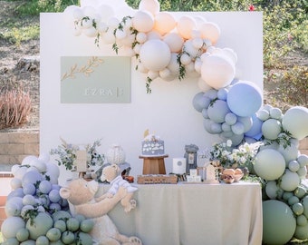 Könnte beinhalten: Ein Babyparty-Setup mit einem weißen Hintergrund, der mit einem Ballonbogen in den Farben Weiß, Pfirsich, Blau und Grün dekoriert ist. Der Tisch ist mit einer Torte, einem Teddybär und anderen Dekorationen gedeckt. Das Schild auf dem Hintergrund sagt "Ezra".