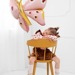 May include: A young girl wearing a brown dress and white socks sits in a yellow chair. A large pink butterfly balloon with red spots is floating above her head.