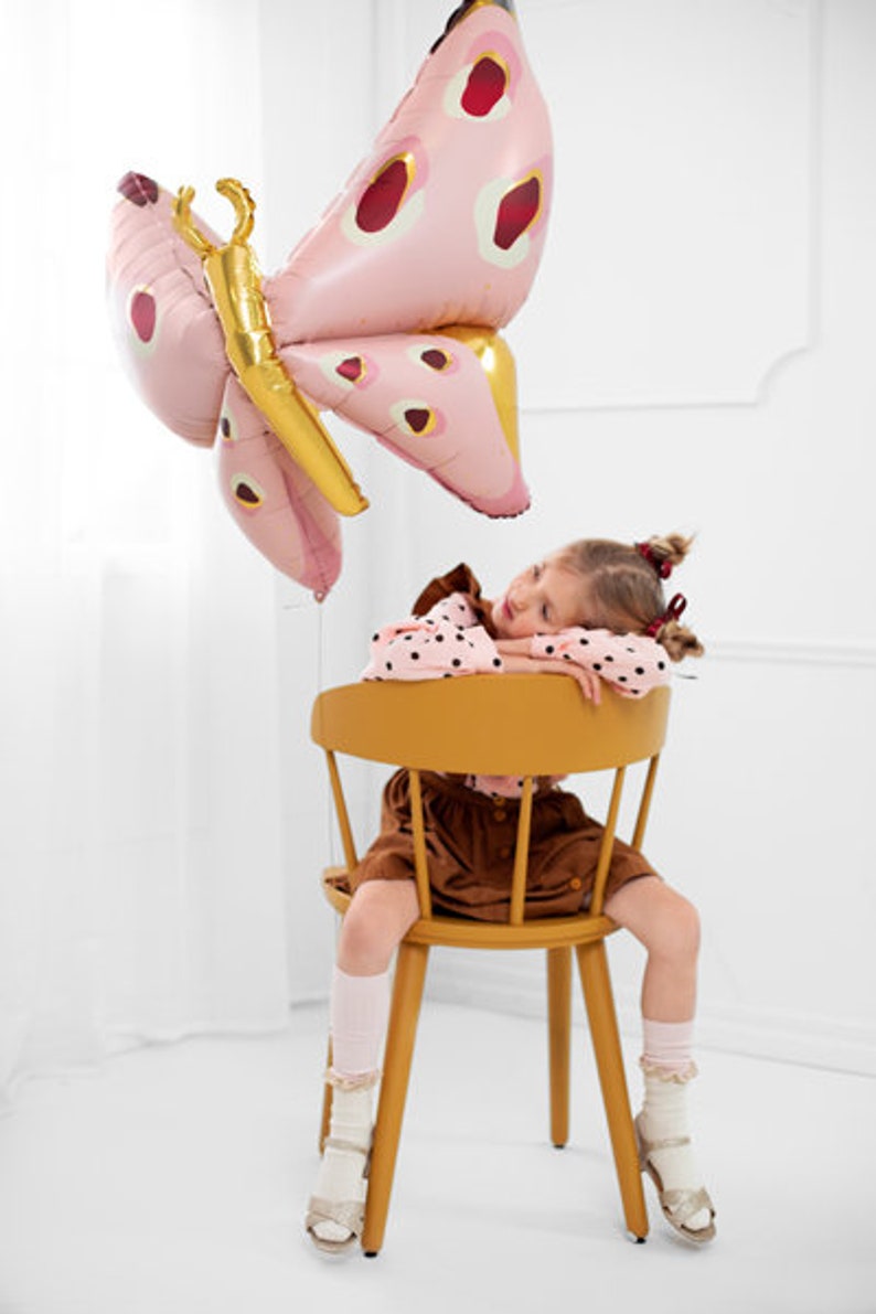May include: A young girl wearing a brown dress and white socks sits in a yellow chair. A large pink butterfly balloon with red spots is floating above her head.