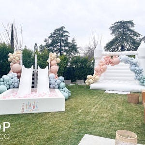 May include: A white inflatable bounce house with a pastel balloon arch and a white inflatable slide with a ball pit. The ball pit has pink balls and a sign that says "Disneyland".