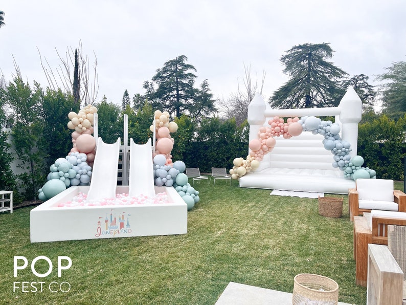 May include: A white inflatable bounce house with a pastel balloon arch and a white inflatable slide with a ball pit. The ball pit has pink balls and a sign that says "Disneyland".