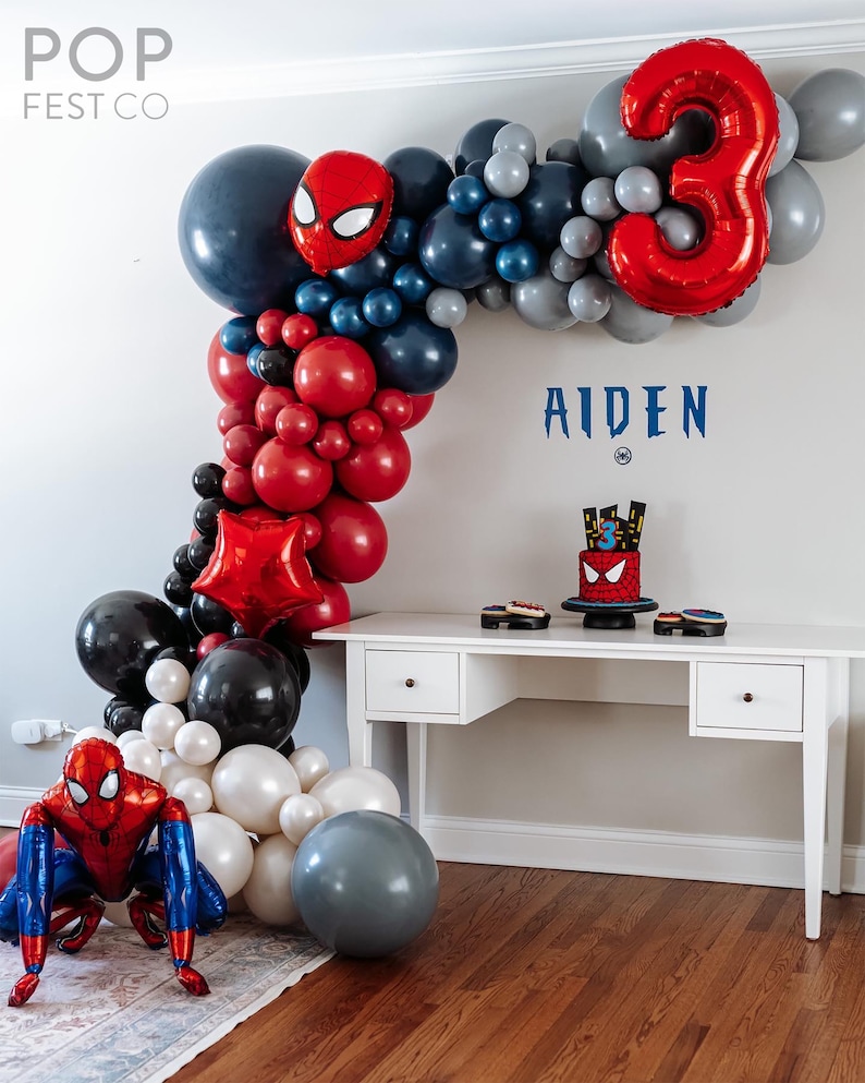 May include: A red, black, and white balloon arch with a Spiderman character balloon and a large red number 3 balloon. The arch is decorated with a Spiderman cake and the name Aiden.