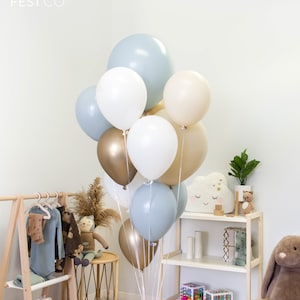 May include: A bouquet of balloons in shades of blue, white, and gold. The balloons are arranged in a cluster and tied together with clear ribbon. The balloons are in a room with a wooden shelf and a wooden rack with clothes hanging on it.