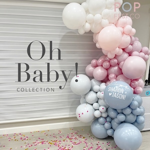 May include: A balloon arch made of white, pink, and blue balloons. The arch is decorated with a sign that says "Sharon + Jason" and the text "Oh Baby! Collection" is on the wall behind the arch.
