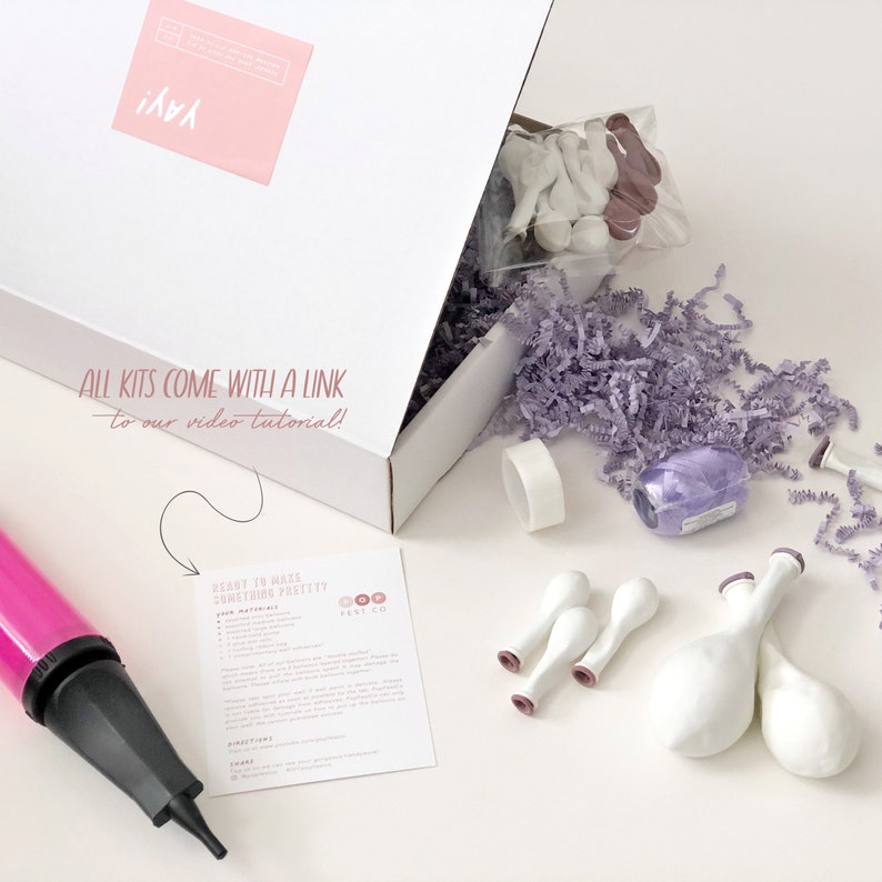 May include: A white box containing a purple and white balloon garland kit. The box is open, revealing the contents: balloons, ribbon, tape, and a hand pump. The box has the text "All kits come with a link to our video tutorial!" printed on the side.