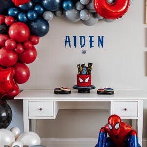 May include: A Spiderman themed birthday party setup with red and blue balloons, a Spiderman cake, and a Spiderman balloon figure. The table is decorated with Spiderman cookies. The name "AIDEN" is written on the wall.