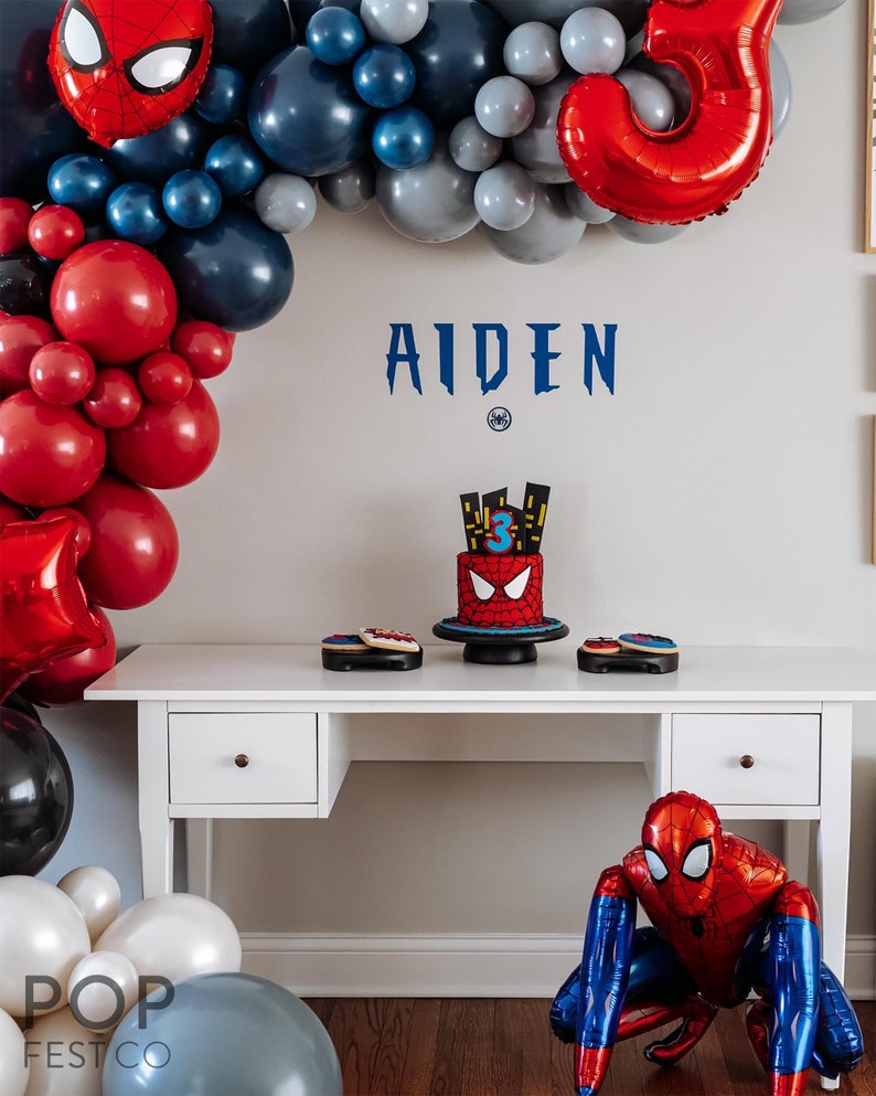 May include: A Spiderman themed birthday party setup with red and blue balloons, a Spiderman cake, and a Spiderman balloon figure. The table is decorated with Spiderman cookies. The name "AIDEN" is written on the wall.