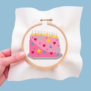 Puede incluir: Bordado de punto de cruz de una tarta de cumpleaños rosa con velas, corazones, estrellas y detalles azules y amarillos. El bordado está colocado en un bastidor de madera y se sostiene sobre un fondo de tela blanca.
