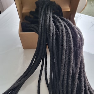 May include: A bundle of dark gray dreadlocks, with a thick, rope-like texture, spilling out of a small, open cardboard box. The dreadlocks are a deep, solid color, and appear to be made of a soft, fibrous material.