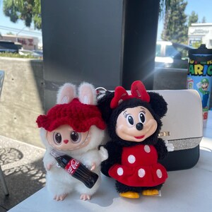 May include: Two plush toys: a white creature with a red hat holding a Coca-Cola bottle, and a Minnie Mouse doll in a red polka-dot dress. A white purse with a black strap is in the background.