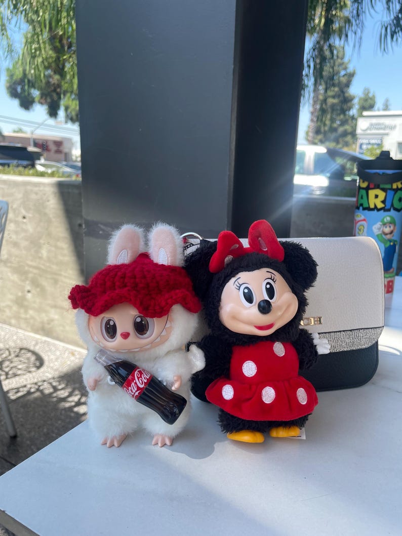 May include: Two plush toys: a white creature with a red hat holding a Coca-Cola bottle, and a Minnie Mouse doll in a red polka-dot dress. A white purse with a black strap is in the background.