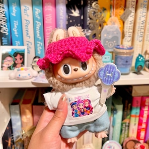 May include: A plush toy with a pink crocheted hat and a white shirt with the word "HUNTRIK" and a graphic of three figures. The toy is holding a blue and white accessory. Bookshelves are in the background.