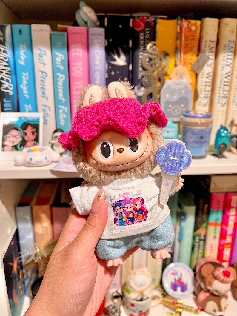 May include: A plush toy with a pink crocheted hat and a white shirt with the word "HUNTRIK" and a graphic of three figures. The toy is holding a blue and white accessory. Bookshelves are in the background.
