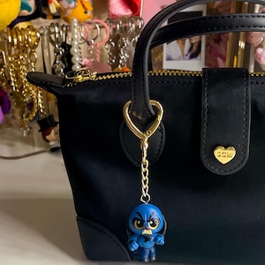 May include: A black handbag with a gold zipper and handle, featuring a gold heart-shaped charm. A blue figurine keychain with a gold chain hangs from the handle. Other keychains are visible in the background.
