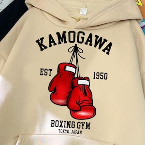 May include: Beige hoodie with a front pocket and a hood. The front features a graphic of two red boxing gloves with the text "KAMOGAWA EST 1950 BOXING GYM TOKYO JAPAN". The hoodie is made of a soft material.