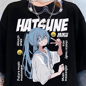 Peut inclure: T-shirt noir avec un graphisme d'un personnage d'anime aux cheveux bleus et le texte "Hatsune Miku". Le texte supplémentaire comprend "Always smile", "Future music messenger" et "Music messenger".