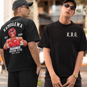 May include: Black t-shirts with boxing-themed graphics. One shirt features "KAMOGAWA BOXING GYM TOKYO JAPAN" with a boxer illustration. The other shirt displays "K.B.G." in white lettering. Both shirts are short-sleeved and have a casual fit.