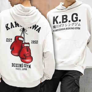 May include: Off-white hoodie with a front pocket and a hood. The back features a graphic of red boxing gloves and the text "KAM...WA EST 1950 BOXING GYM TOKYO, JAPAN." The front displays "K.B.G. 鴨川ボクシングジム KAMOGAWA BOXING GYM."
