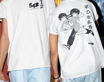 Könnte beinhalten: Weiße T-Shirts mit japanischem Text und Illustrationen im Anime-Stil. Ein Shirt hat Text und ein Logo auf der Vorderseite, das andere eine Grafik auf der Rückseite. Die Shirts werden mit hellblauen Jeans getragen.