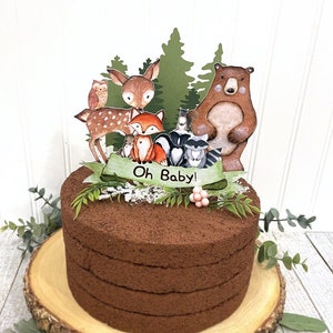 May include: A chocolate cake with a woodland animal themed cake topper. The topper features a deer, a bear, a fox, an owl, and a raccoon. The banner reads "Oh Baby!"
