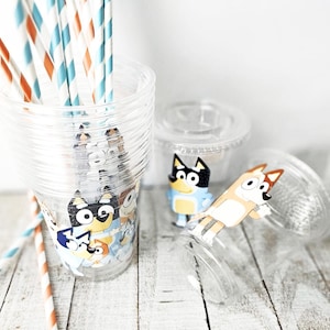 May include: Clear plastic cups with cartoon character designs, stacked and scattered on a white wooden surface. The cups are decorated with colorful cartoon characters. Striped straws in orange, blue, and white are in one cup.