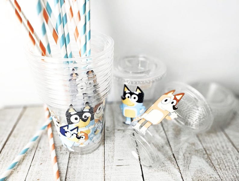 May include: Clear plastic cups with cartoon character designs, stacked and scattered on a white wooden surface. The cups are decorated with colorful cartoon characters. Striped straws in orange, blue, and white are in one cup.