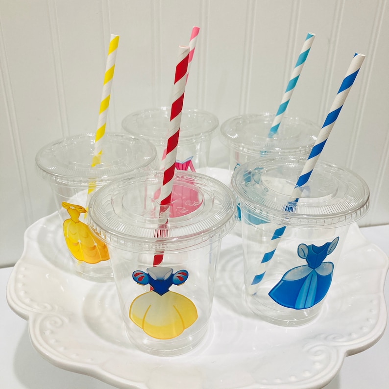May include: Six clear plastic cups with colorful straws and princess-themed designs. The cups have lids and feature images of princesses in different colored dresses.