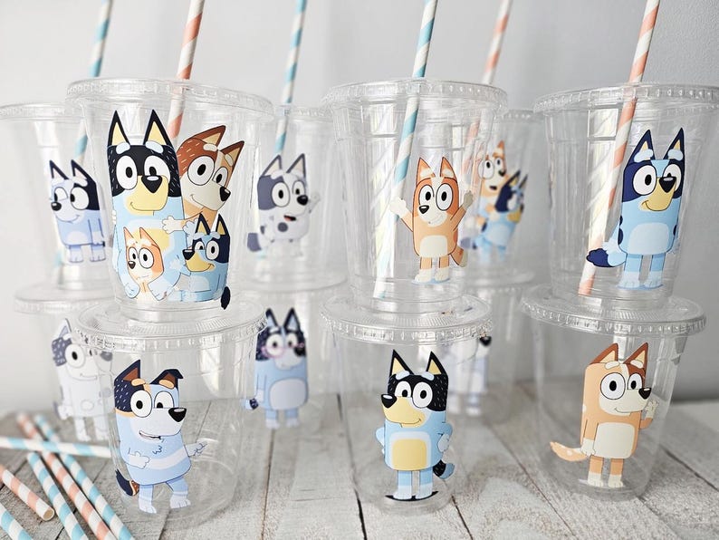 May include: Clear plastic cups with lids and straws, decorated with cartoon dog characters in various poses. The straws have blue and pink stripes. The cups are stacked on top of each other.