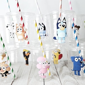 May include: Clear plastic cups with lids and colorful paper straws. Each cup features a different cartoon dog character sticker. Straws have red, green, blue, and yellow stripes. The cups are arranged on a light-colored wooden surface.