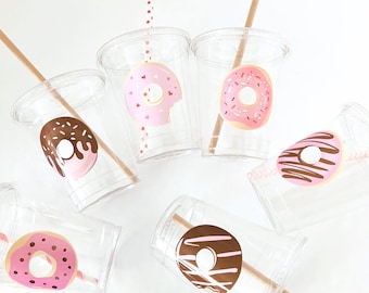 May include: Clear plastic cups with donut designs in pink and brown, each with a straw. Some donuts have sprinkles, and one has a bite taken out. The straws are brown and red and white striped.
