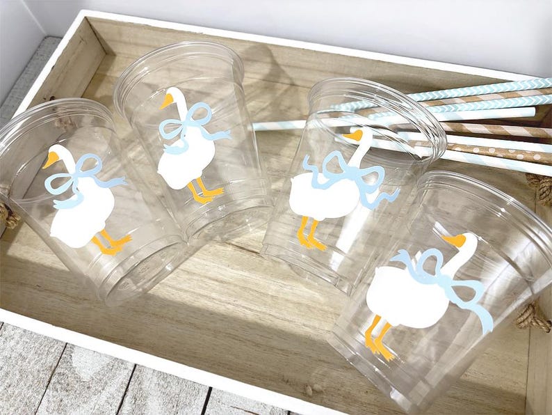 May include: Four clear plastic cups with a white goose design, each with a light blue bow and yellow feet. The cups are in a wooden tray with striped straws.