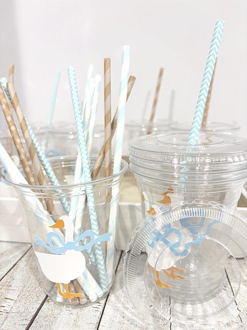 May include: Clear plastic cups with lids and paper straws. The cups are decorated with a white goose wearing a blue bow. The straws are brown, white, and light blue. The cups are on a light wood surface.