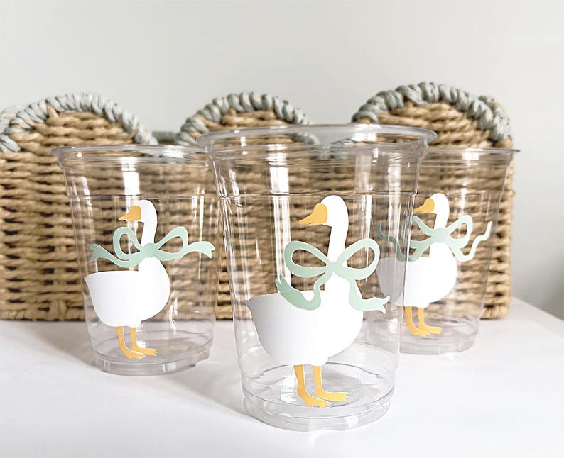 May include: Clear plastic cups with a white goose design, featuring a yellow beak and feet, and a light green bow. The cups are in front of a woven basket, suggesting a party or event theme.