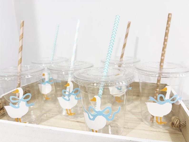 May include: Six clear plastic cups with blue and white duck designs and blue and white striped straws. The cups are filled with a clear liquid. The cups are on a white surface.