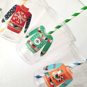 May include: Three clear plastic cups with festive holiday sweater designs. Each cup has a different design featuring a snowflake, geometric patterns, and a snowman. The cups are decorated with colorful patterns and have a straw.