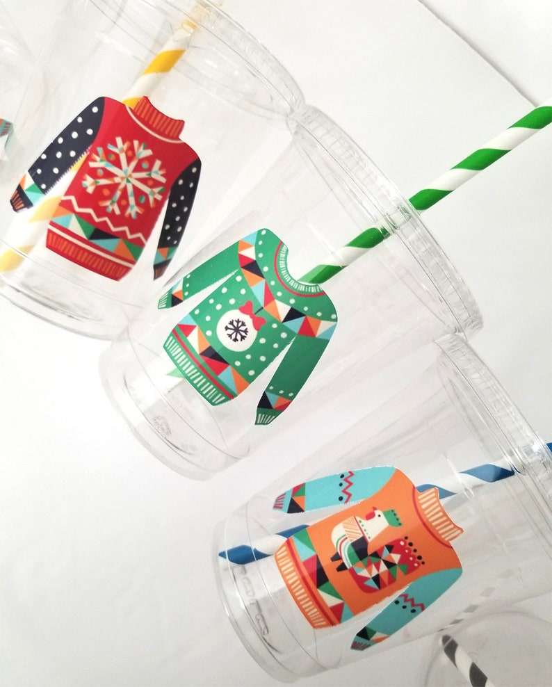 May include: Three clear plastic cups with festive holiday sweater designs. Each cup has a different design featuring a snowflake, geometric patterns, and a snowman. The cups are decorated with colorful patterns and have a straw.