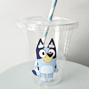 May include: A clear plastic cup with a lid and a blue and white striped straw. The cup features a cartoon image of a blue dog with black and white accents. The cup is designed for cold beverages.