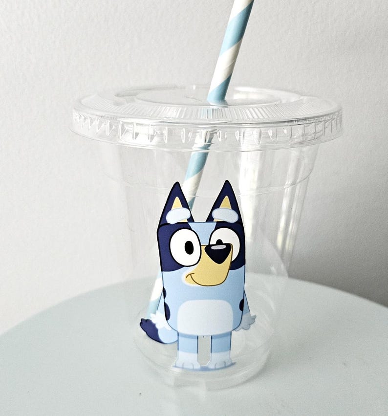 May include: A clear plastic cup with a lid and a blue and white striped straw. The cup features a cartoon image of a blue dog with black and white accents. The cup is designed for cold beverages.