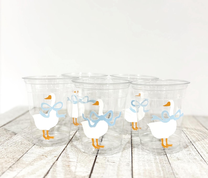 May include: Clear plastic cups with a white goose design, featuring orange beaks and feet, and a light blue bow. The cups are arranged on a light wood surface. The cups are suitable for a baby shower or a children's party.