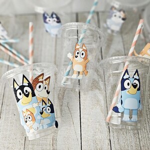 May include: Clear plastic cups with Bluey cartoon character designs. Each cup has a straw with blue and peach stripes. The cups are on a light wood surface, with additional Bluey character cutouts scattered around.