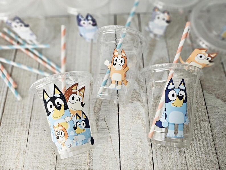 May include: Clear plastic cups with Bluey cartoon character designs. Each cup has a straw with blue and peach stripes. The cups are on a light wood surface, with additional Bluey character cutouts scattered around.